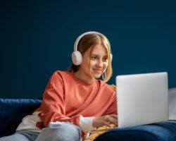 Smiling teenage girl wearing headphones typing on laptop engaging in online teaching