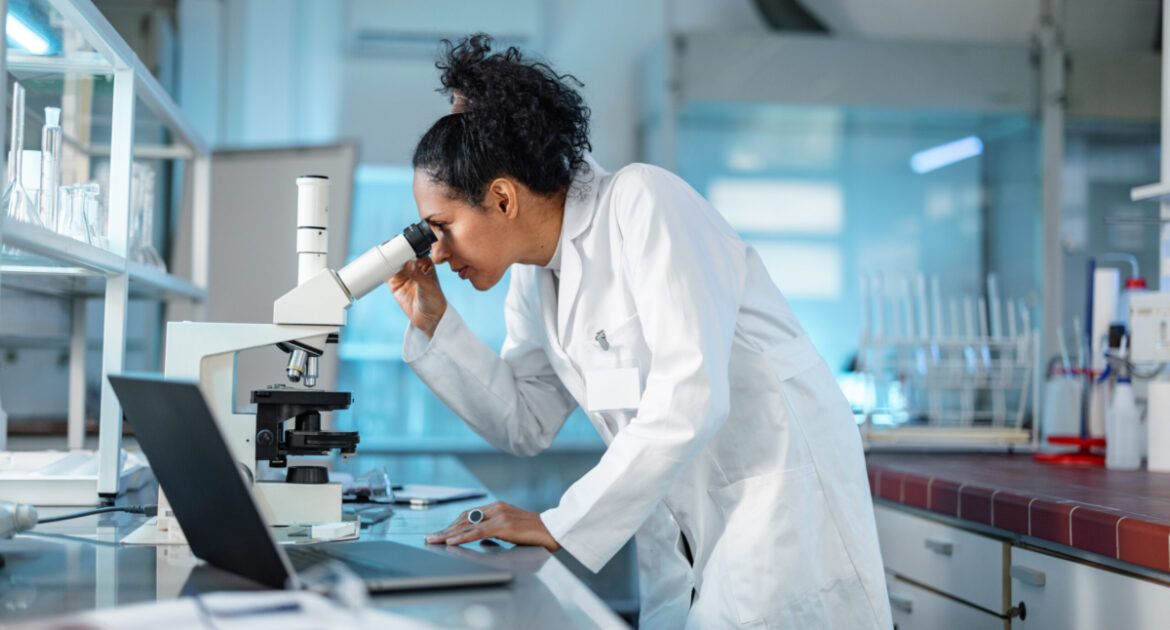Female Scientist Looking Under Microscope And Using Laptop In A Laboratory